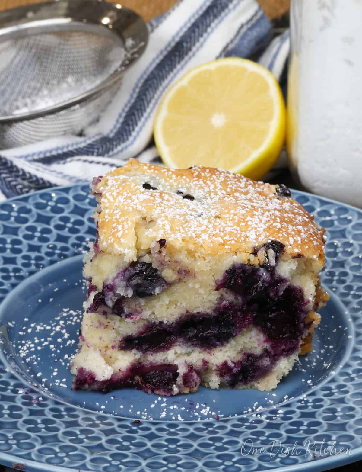 a slice of blueberry cake on a blue plate next to a half of a lemon and a bowl of powdered sugar.
