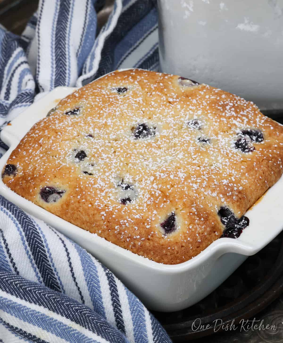 a small blueberry cake in a white baking dish on a silver tray next to a blue and white napkin.
