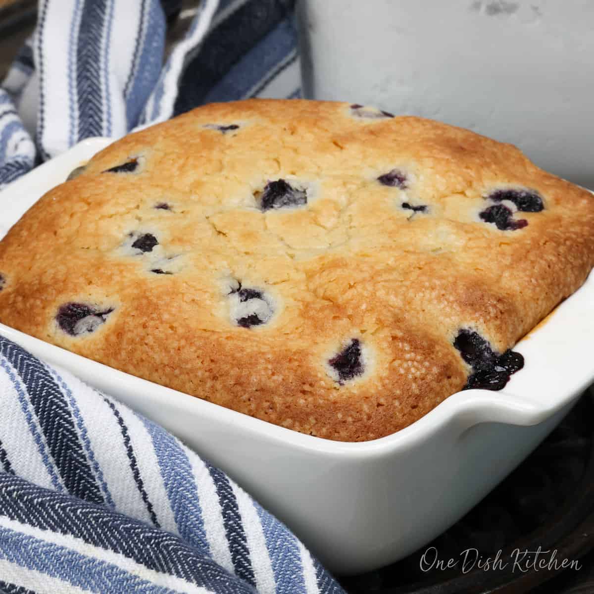 a square cake with blueberries next to a blue towel.