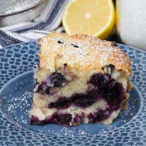 a slice of blueberry cake on a blue plate next to a half of a lemon and a bowl of powdered sugar