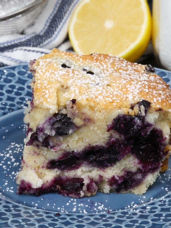 a slice of blueberry cake on a blue plate next to a half of a lemon and a bowl of powdered sugar