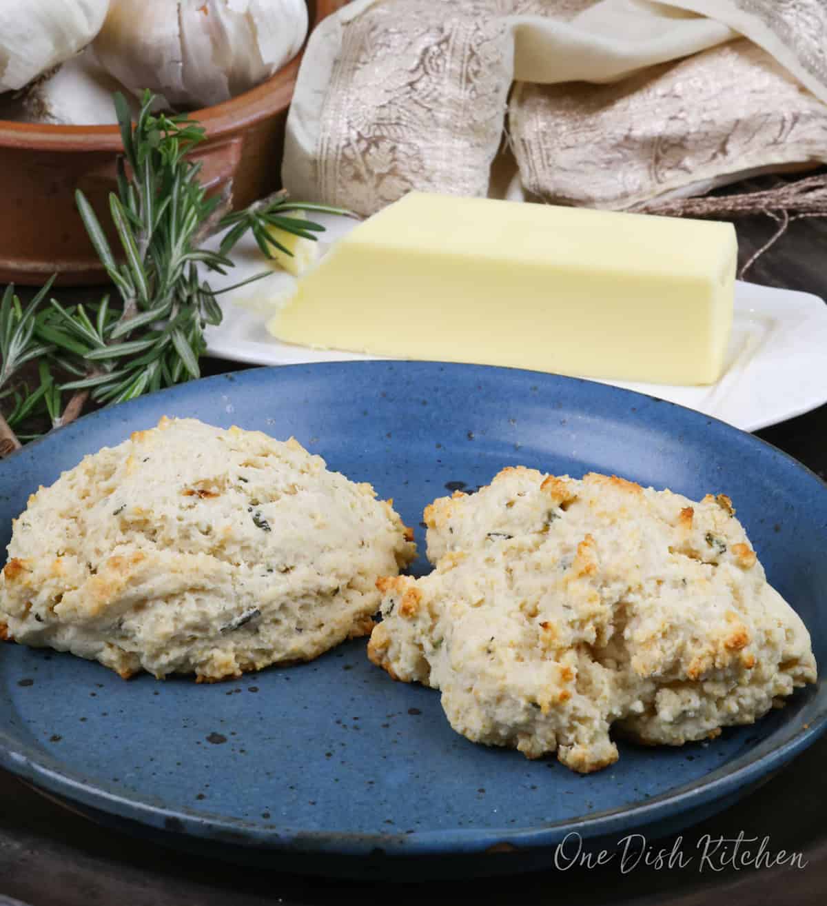 two buttermilk biscuits on a blue plate next to a sprig of fresh rosemary and butter.