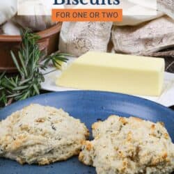 two buttermilk biscuits on a blue plate next to a stick of butter and fresh rosemary.