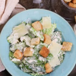 an overhead shot of a blue bowl filled with caesar salad topped with parmesan cheese and croutons next to a jar of homemade caesar dressing