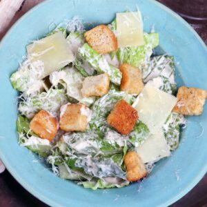 a blue bowl filled with caesar salad topped with parmesan cheese and croutons.