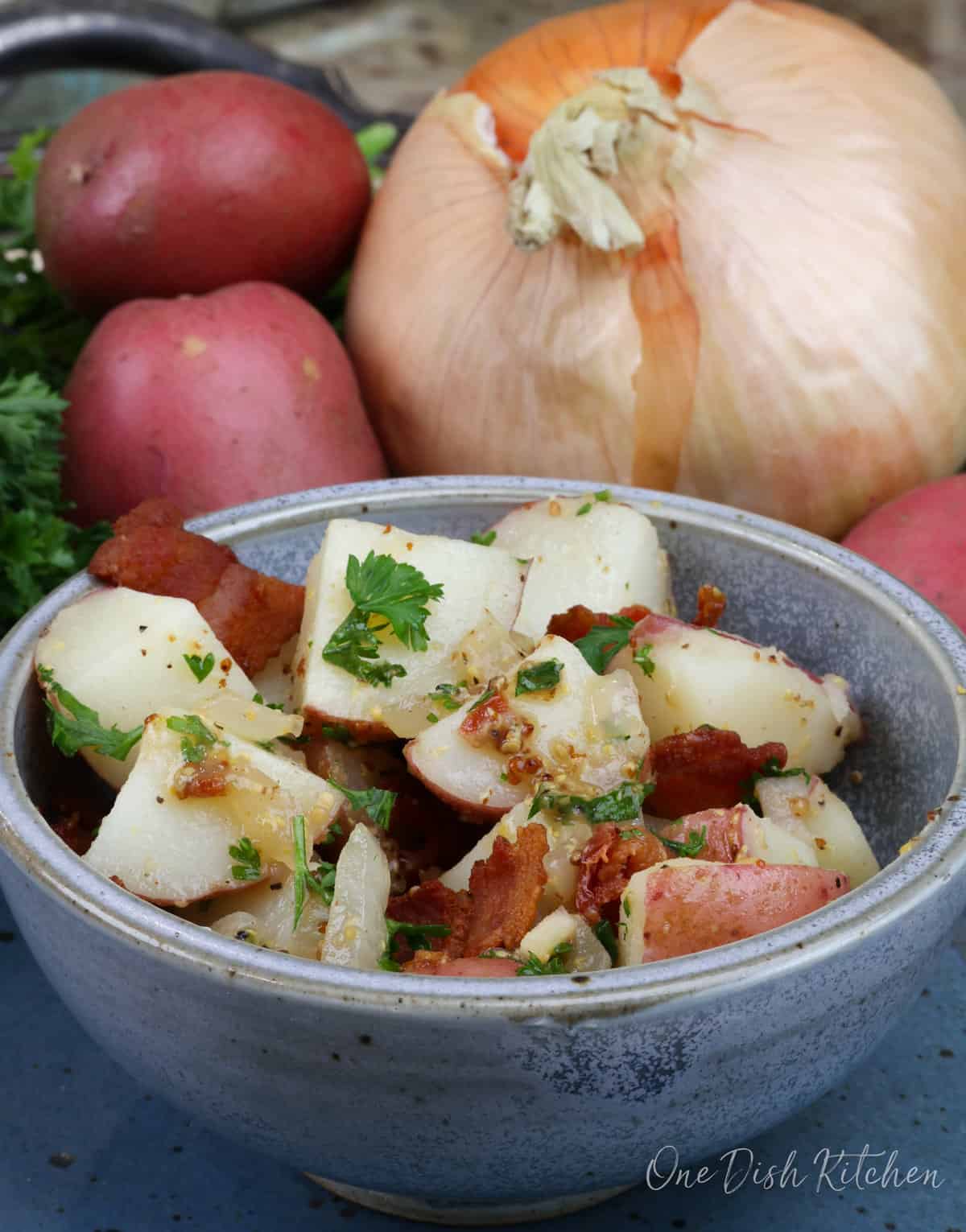 a blue bowl filled with a small batch of german potato salad next to 2 red potatoes and an onion.