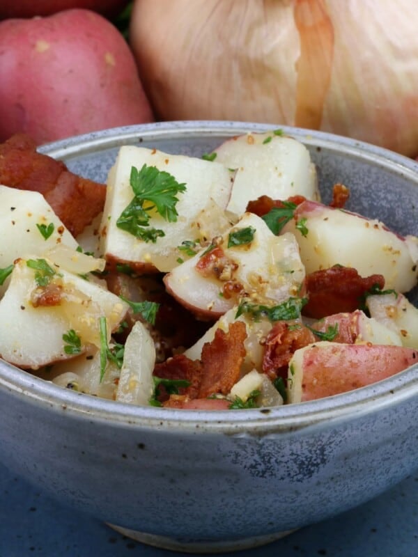 a blue bowl filled with a small batch of german potato salad next to 2 red potatoes and an onion