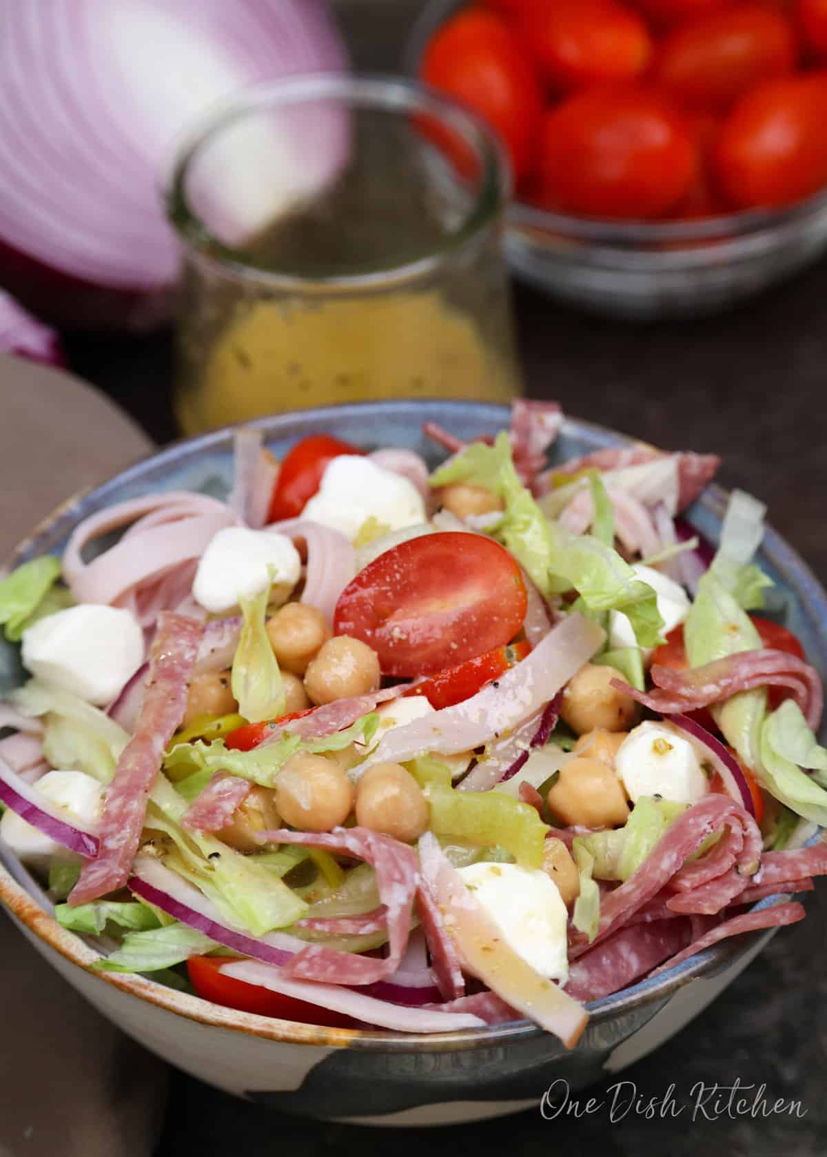 a chopped salad topped with italian meats on a silver tray next to a homemade vinaigrette in a jar.