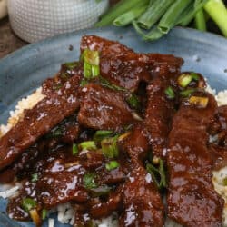 mongolian beef over rice on a blue plate next to fresh green onions
