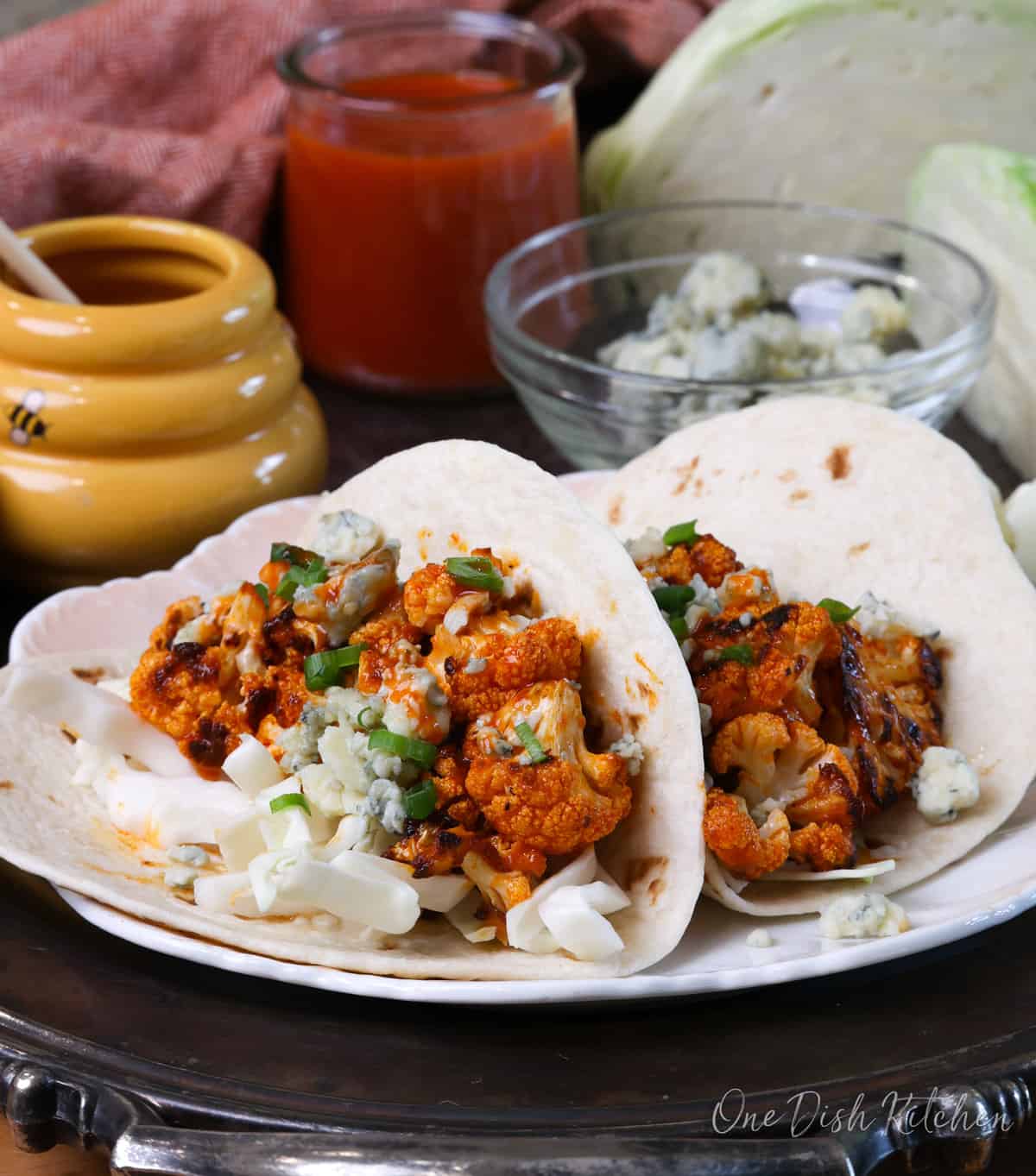 two buffalo cauliflower tacos on a plate next to a bowl of blue cheese crumbles and a jar of honey.