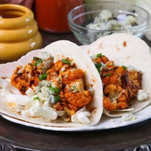 two buffalo cauliflower tacos on a plate topped with buffalo sauce, blue cheese, and shredded cabbage.