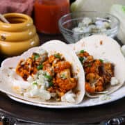 two buffalo cauliflower tacos on a plate next to a bowl of blue cheese crumbles and a jar of honey