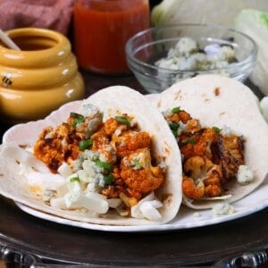 two buffalo cauliflower tacos on a plate next to a bowl of blue cheese crumbles and a jar of honey