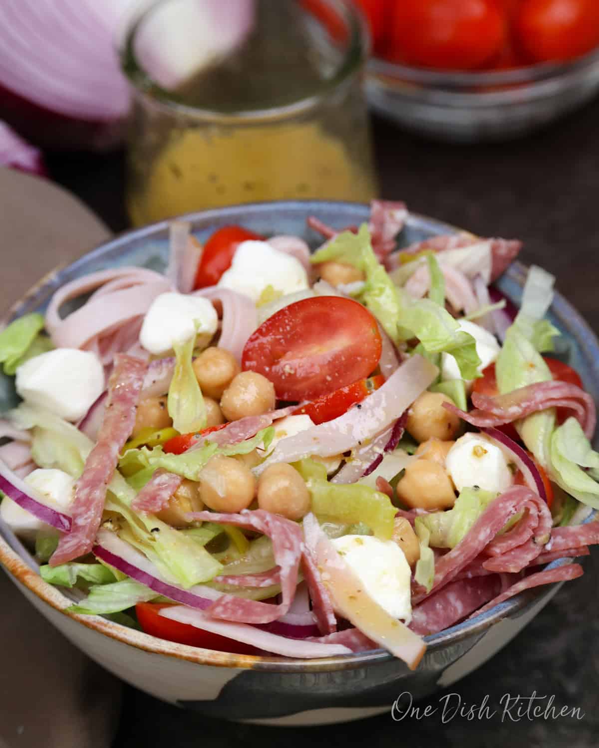 a chopped salad topped with italian meats on a silver tray next to a homemade vinaigrette in a jar.