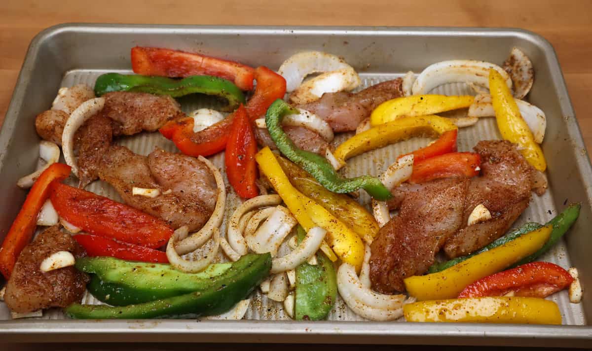 seasoned chicken strips and slices of onions and bell peppers on a sheet pan.