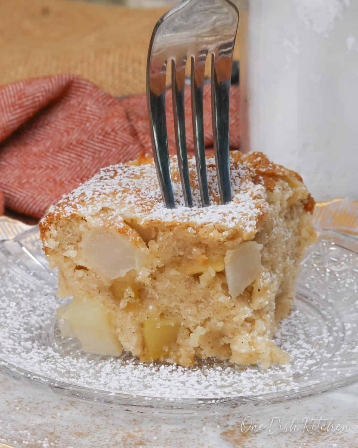 a slice of apple cake topped with powdered sugar on a white plate next to an orange napkin.