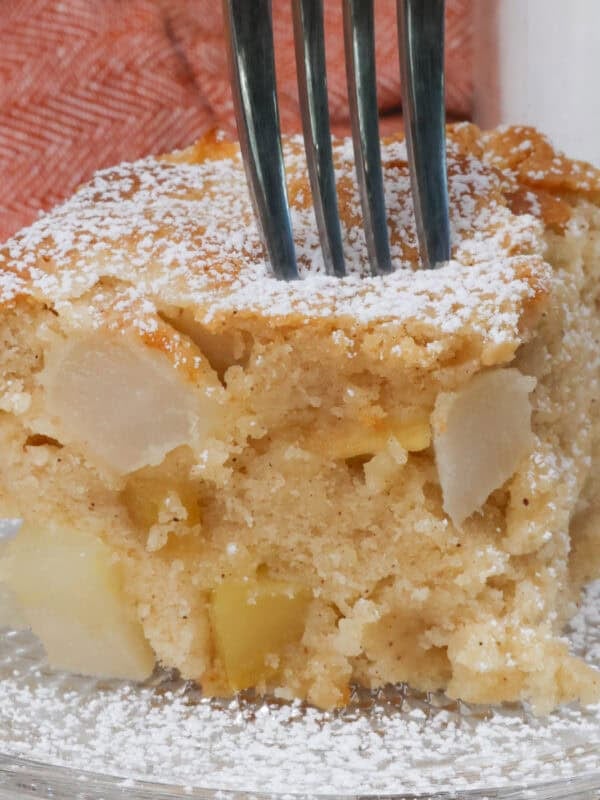 a slice of apple cake topped with powdered sugar on a white plate next to an orange napkin.