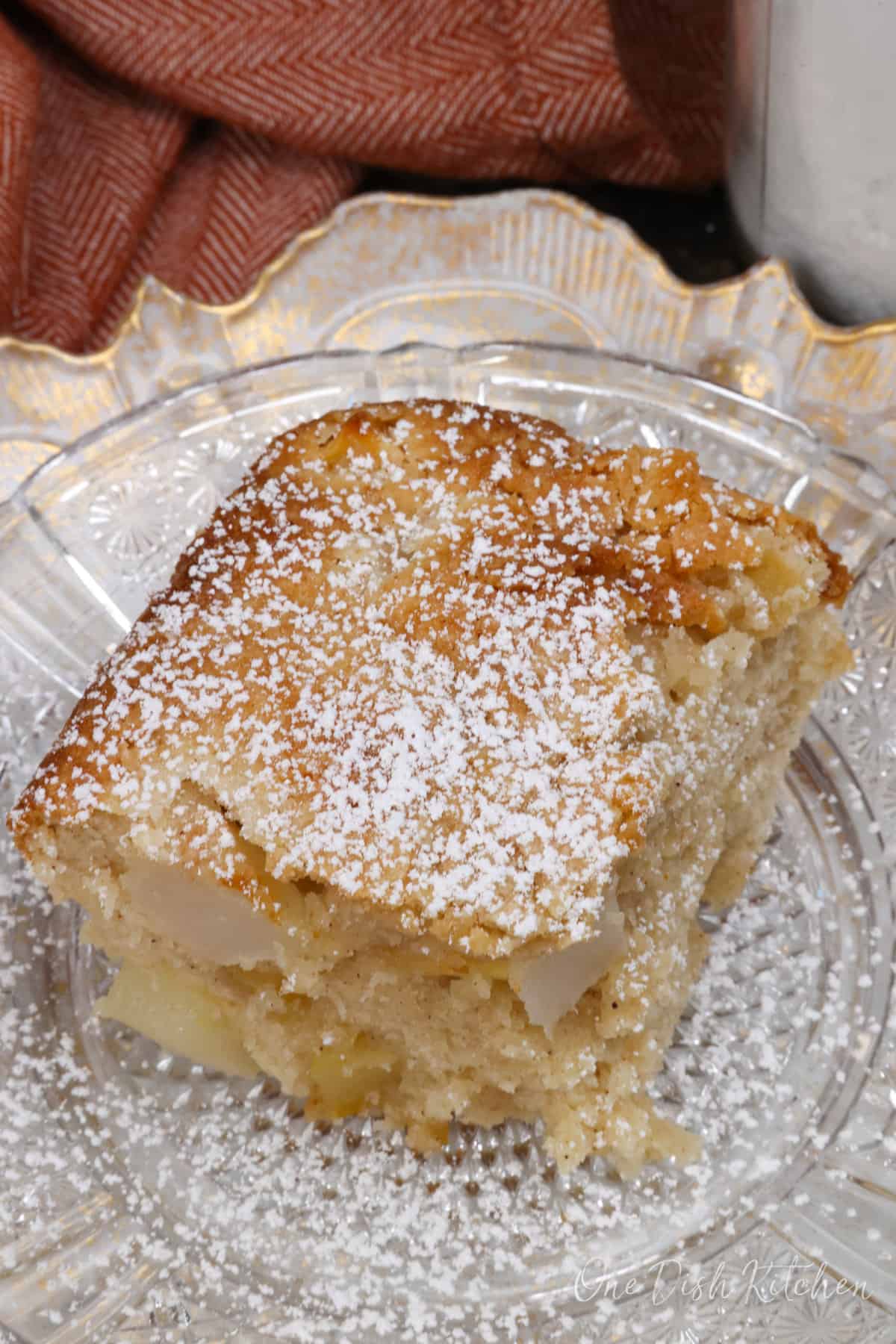 a slice of apple cake topped with powdered sugar on a white plate next to an orange napkin.