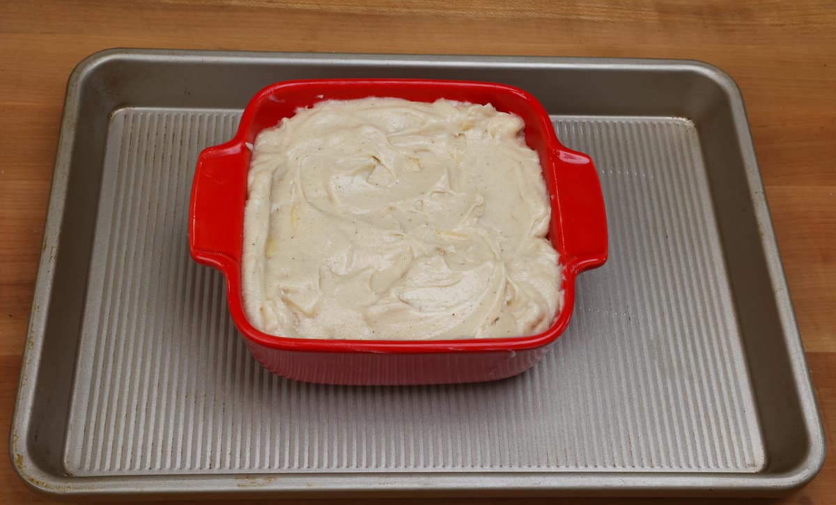 apple cake batter in a square red baking dish on a rimmed baking sheet.