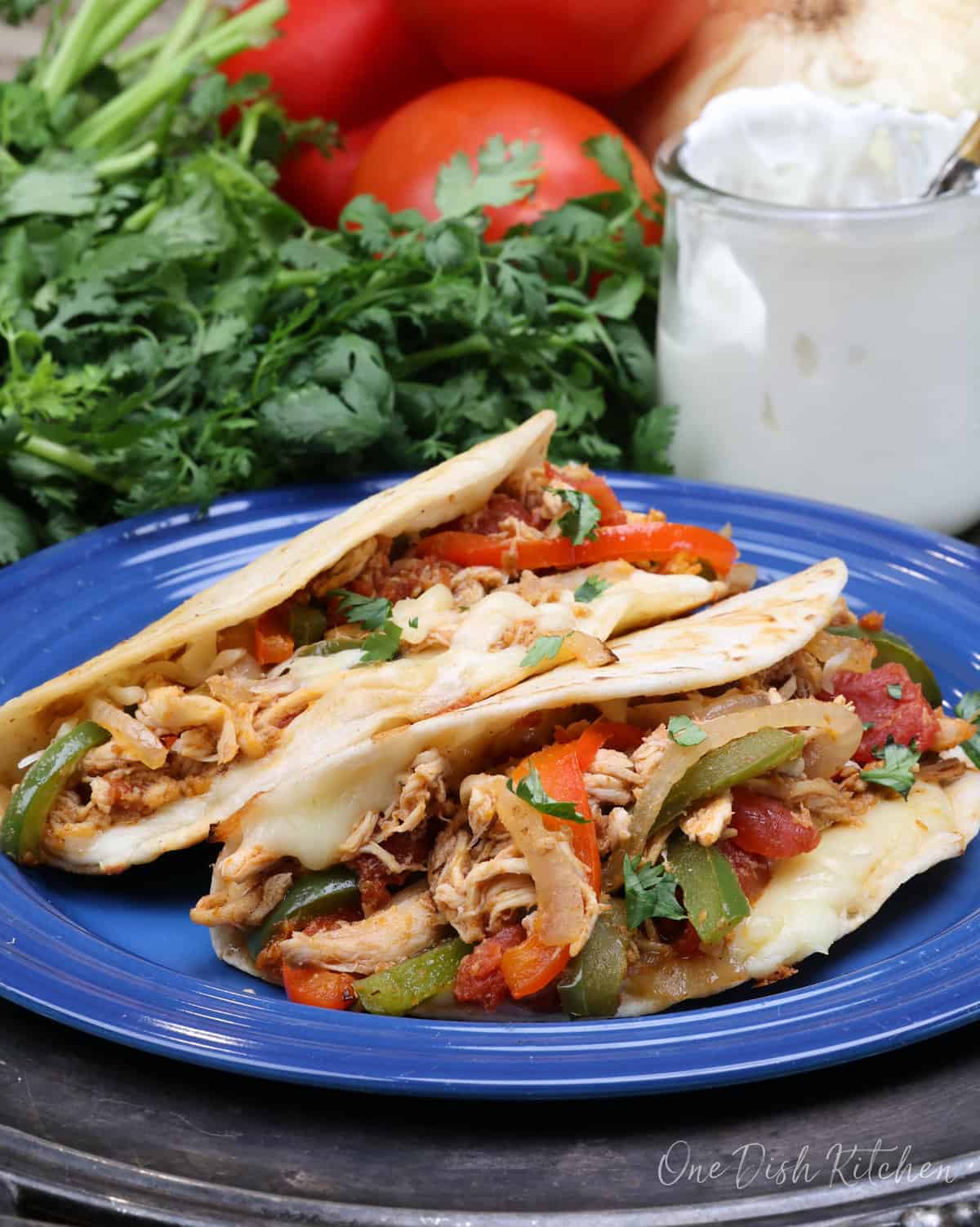 Two chicken tacos on a blue plate surrounded by cilantro, tomatoes, and a jar of sour cream.