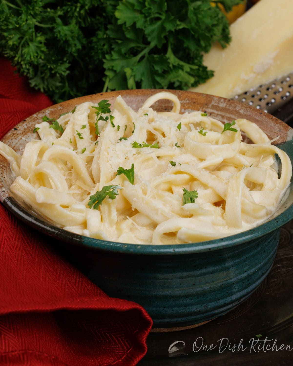 a bowl of fettuccine alfredo topped with chopped parsley next to a red napkin and a wedge of parmesan cheese.