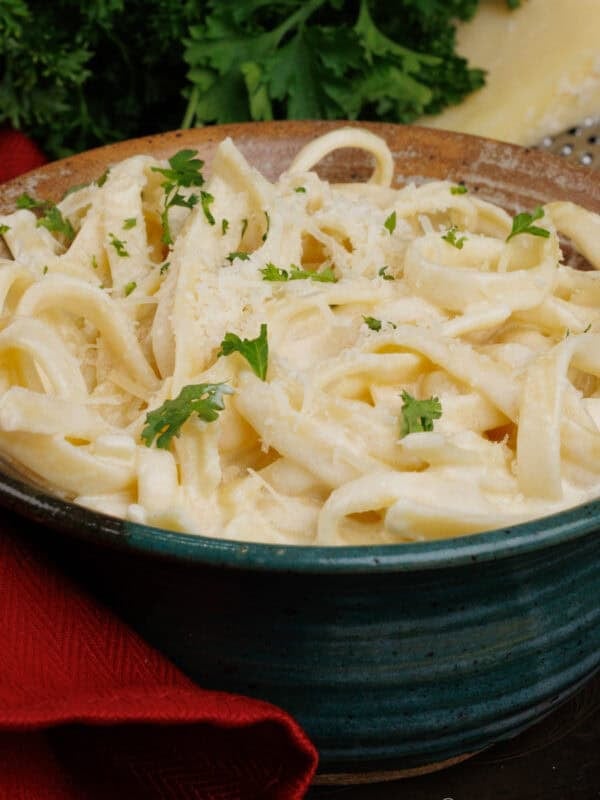 a bowl of fettuccine alfredo topped with chopped parsley next to a red napkin and a wedge of parmesan cheese.