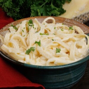 a bowl of fettuccine alfredo topped with chopped parsley next to a red napkin and a wedge of parmesan cheese.