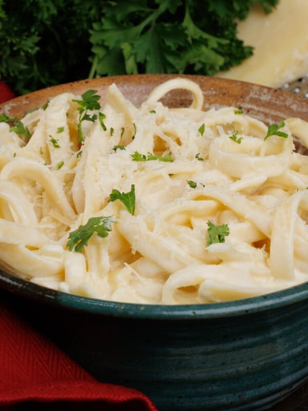 a bowl of fettuccine alfredo topped with chopped parsley next to a red napkin and a wedge of parmesan cheese.