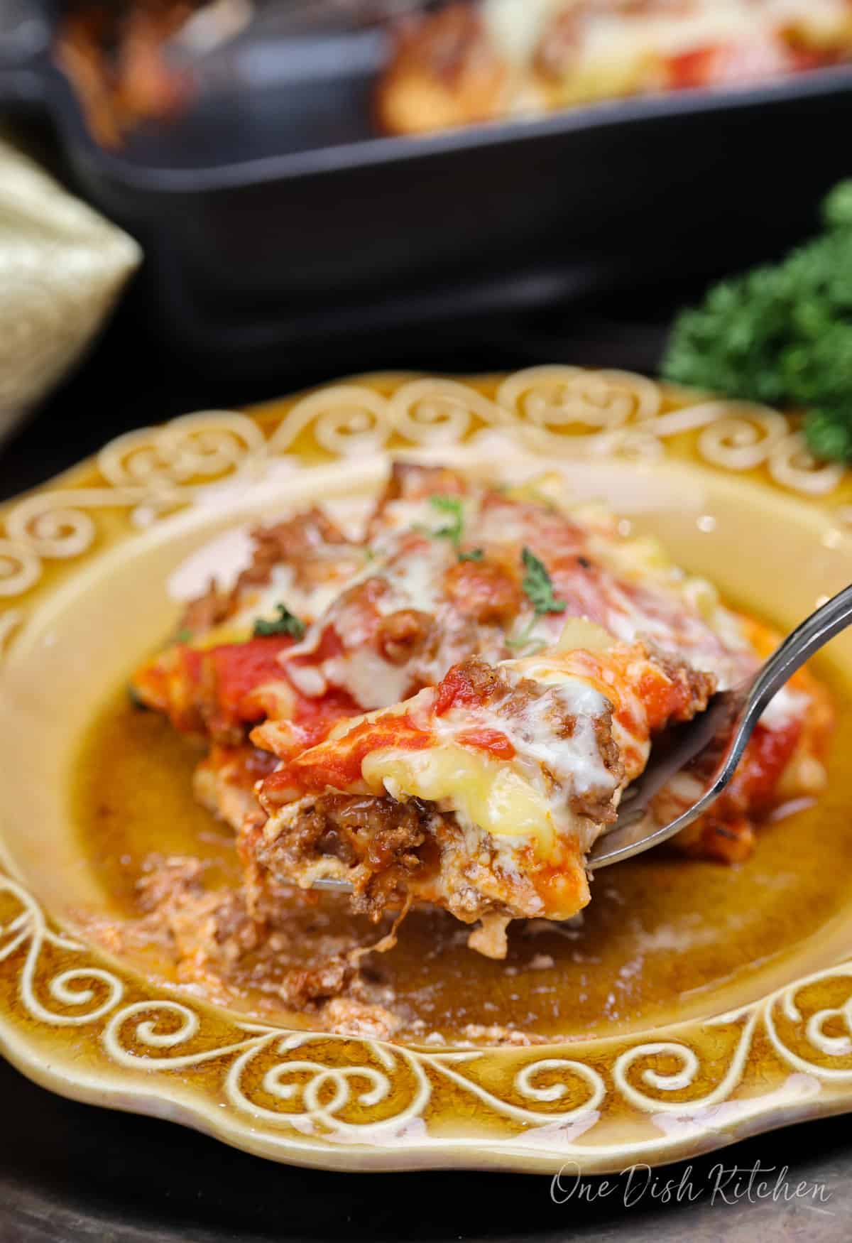 a fork holding lasagna over a gold plate with the remaining serving of lasagna.