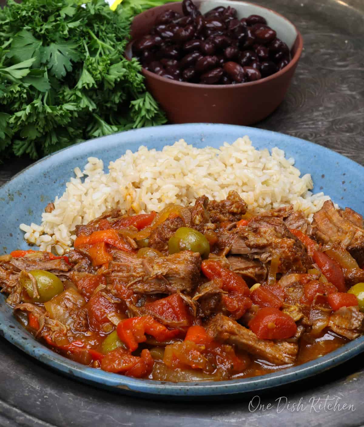 Ropa vieja with rice on a blue plate next to a bowl of black beans.
