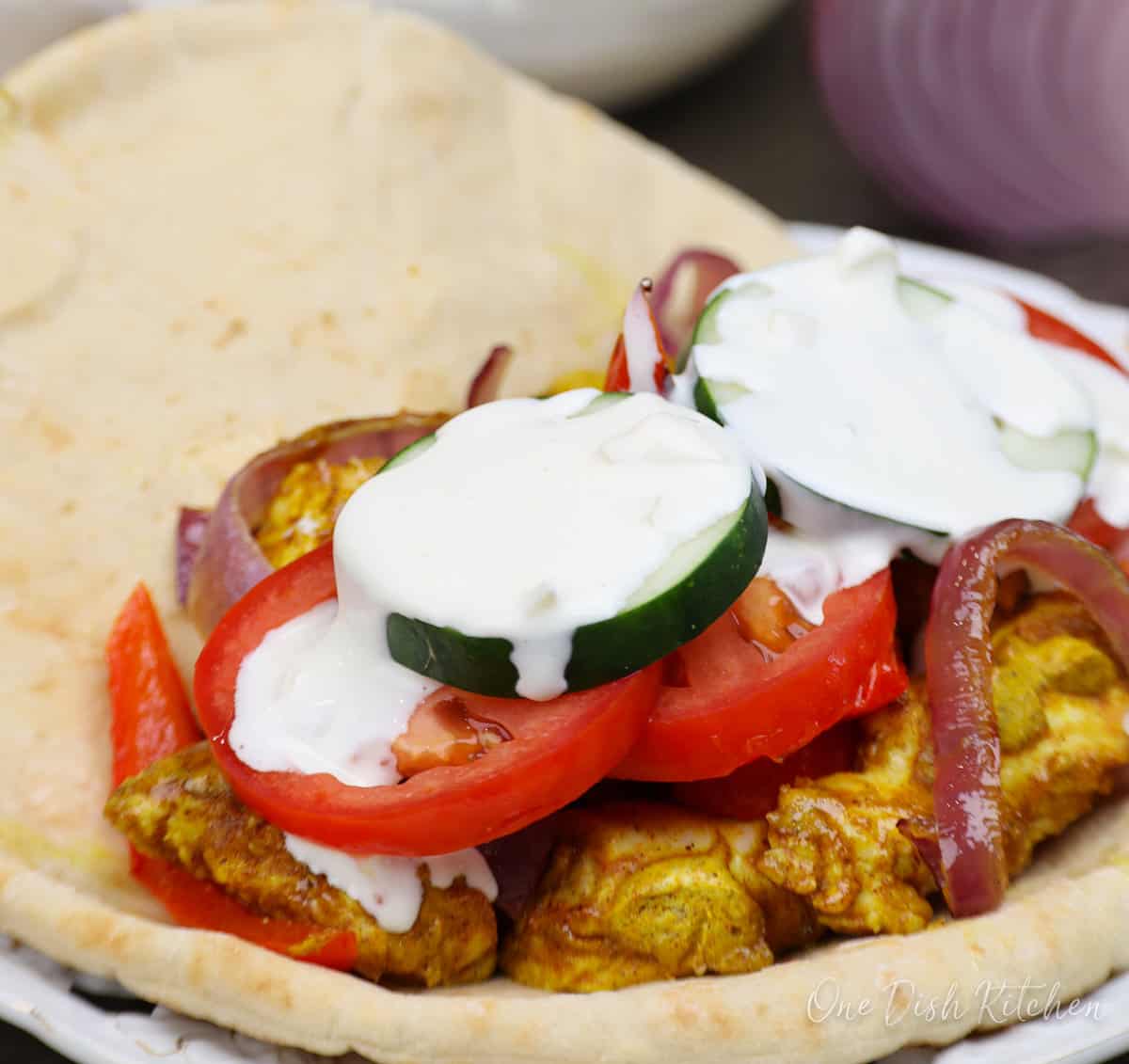 an open-faced chicken shawarma on a white plate topped with white yogurt sauce.