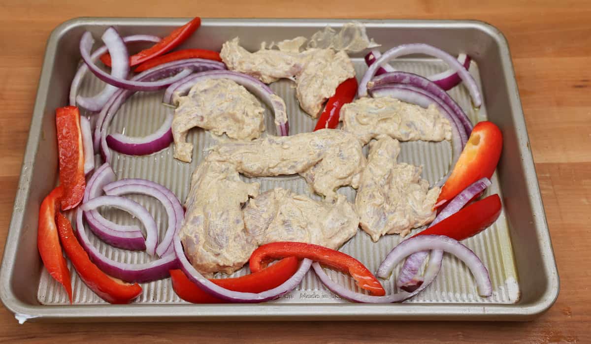 yogurt marinated chicken surrounded by slices of red onion and red bell peppers on a sheet pan.