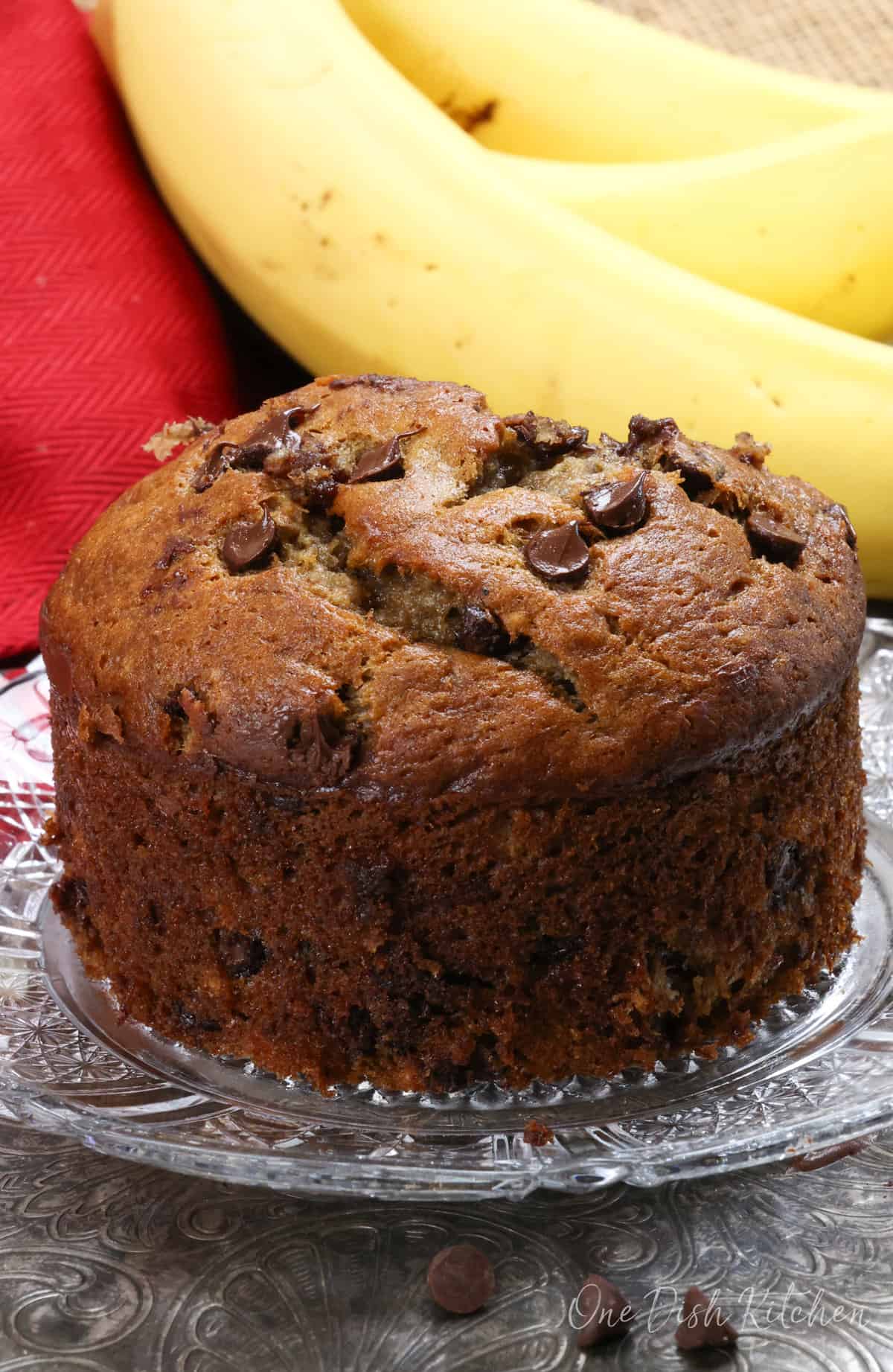 one small chocolate chip banana bread on a plate next to a red napkin and one banana.