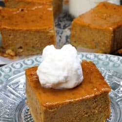 a pumpkin pie bar topped with whipped cream on a plate next to three pumpkin pie bars and a jar or whipped cream.