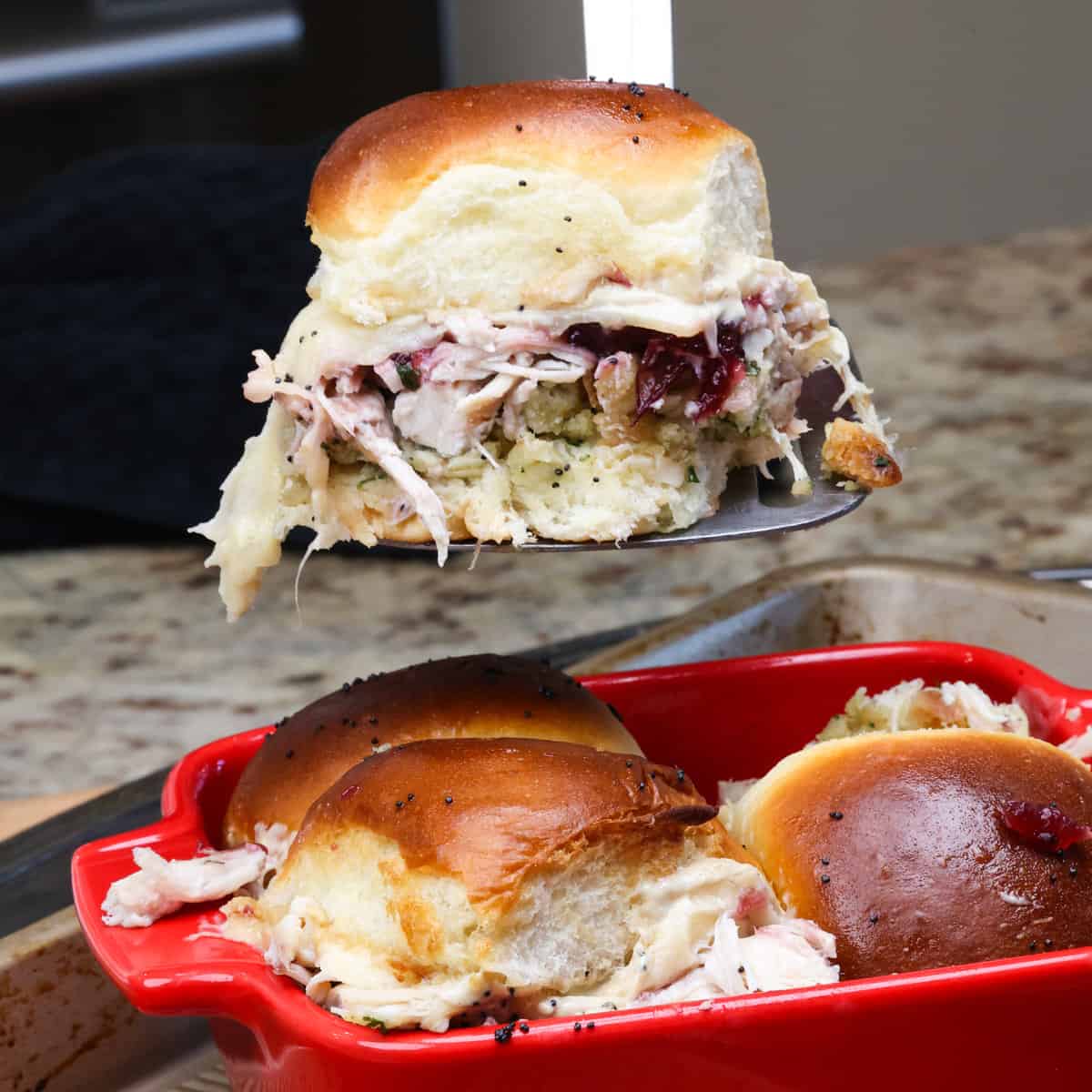 a thanksgiving slider on a metal spatula over a baking dish filled with more sliders.
