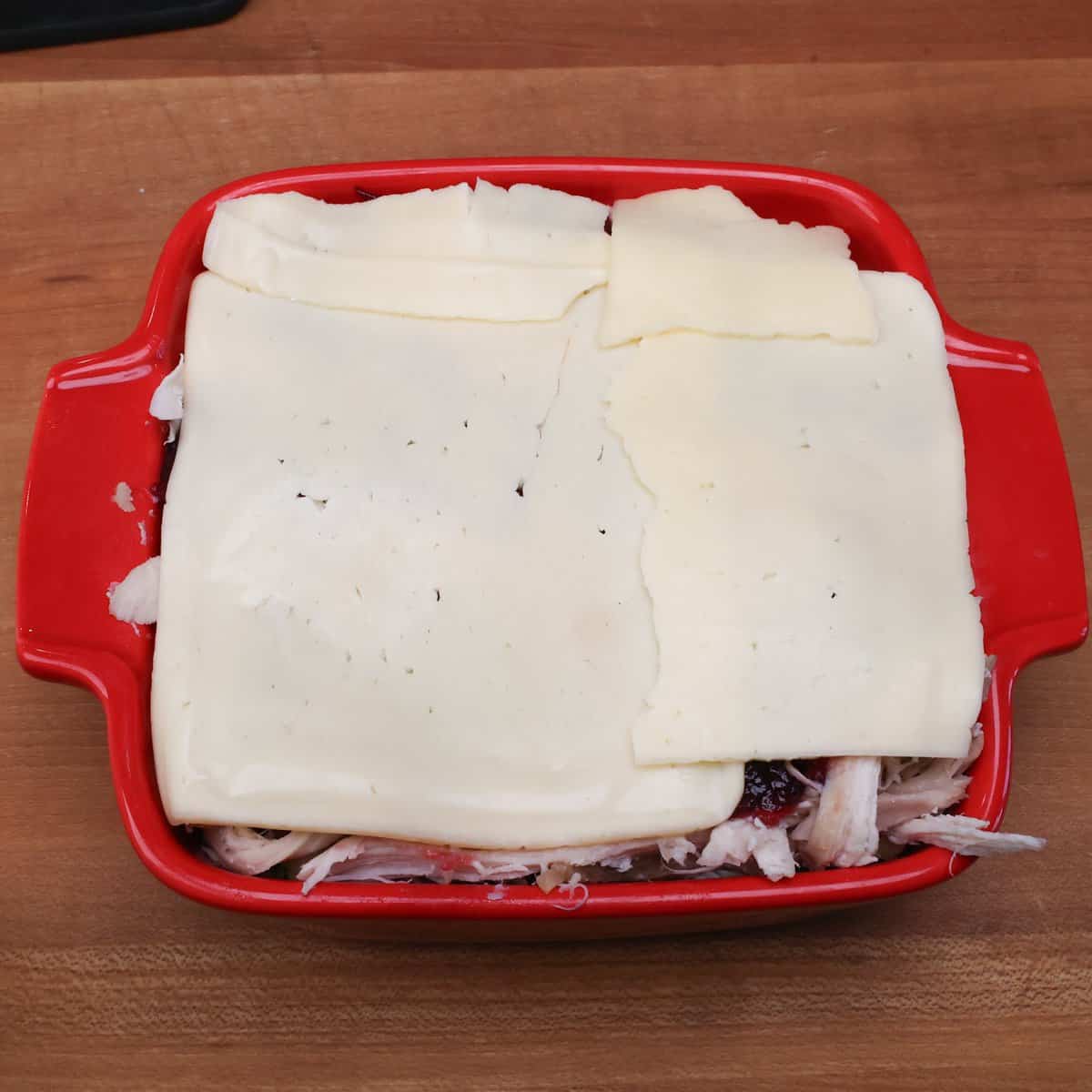 slices of cheese on top of turkey, stuffing, and cranberry sauce in a baking dish.