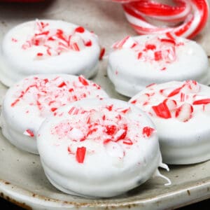 5 oreos coated in white chocolate and topped with candy cane sprinkles on a plate next to a red napkin.