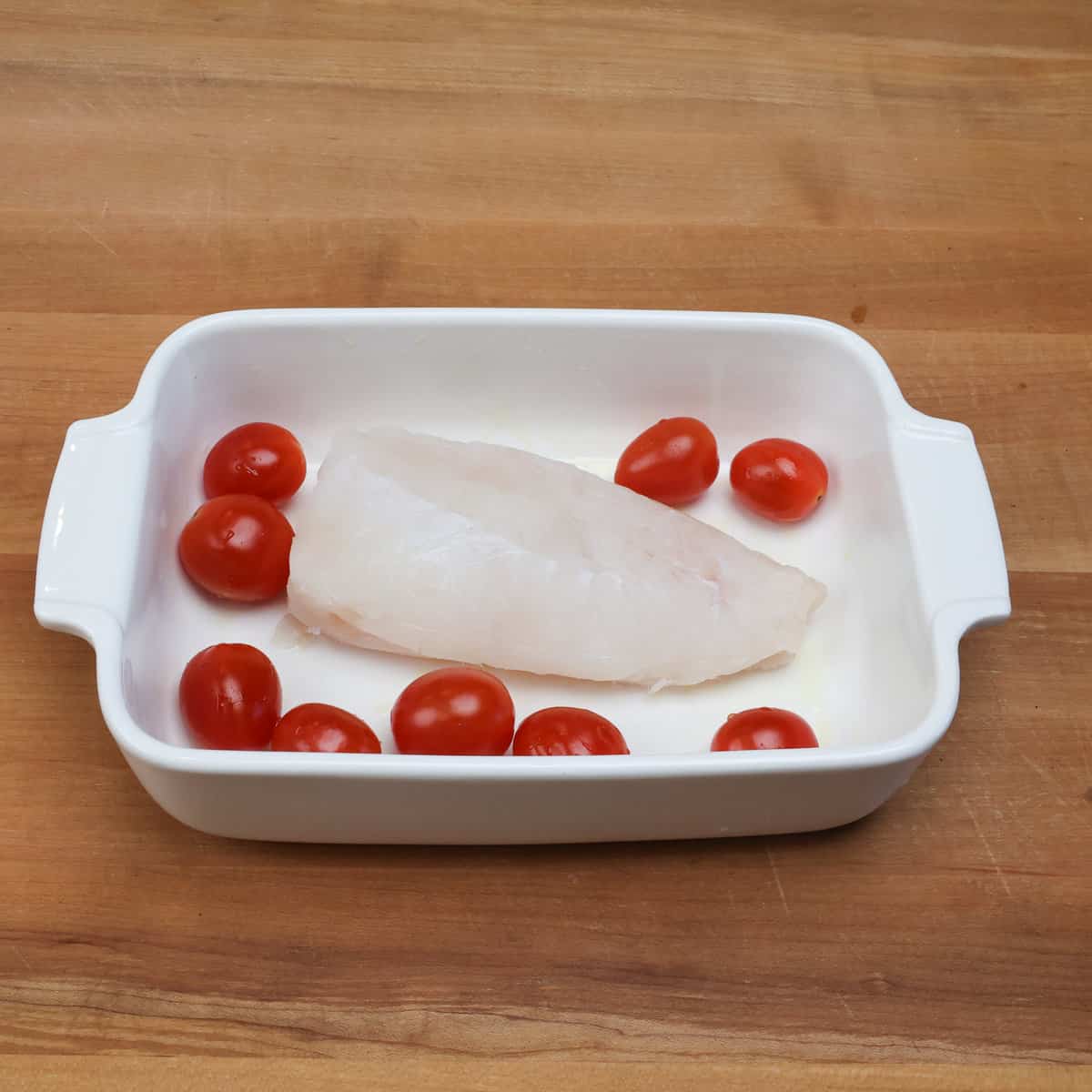 a cod fillet in a white baking dish with cherry tomatoes scattered around the fish.