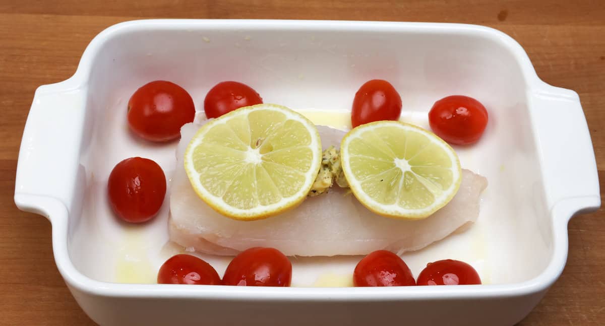Unbaked cod topped with butter and seasonings surrounded by tomatoes and lemons.