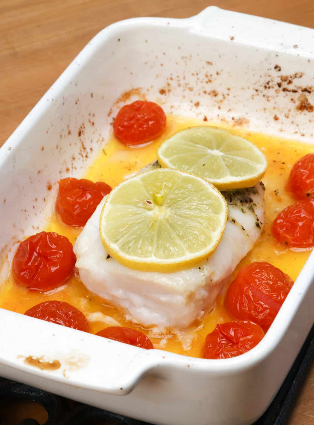 an oven baked cod fillet topped with butter and lemons and surrounded by cherry tomatoes in a baking dish.
