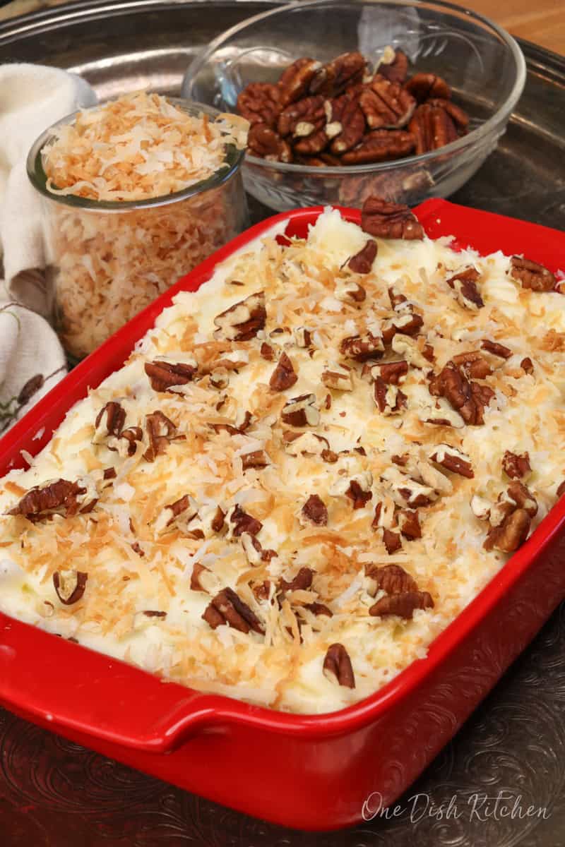 an italian cream cake in a small red square baking dish with toasted shredded coconut and nuts on top.This mini Italian Cream Cake is rich, moist, and filled with toasted pecans and coconut. Topped with a creamy frosting, it’s a classic Southern dessert scaled down for one or two.