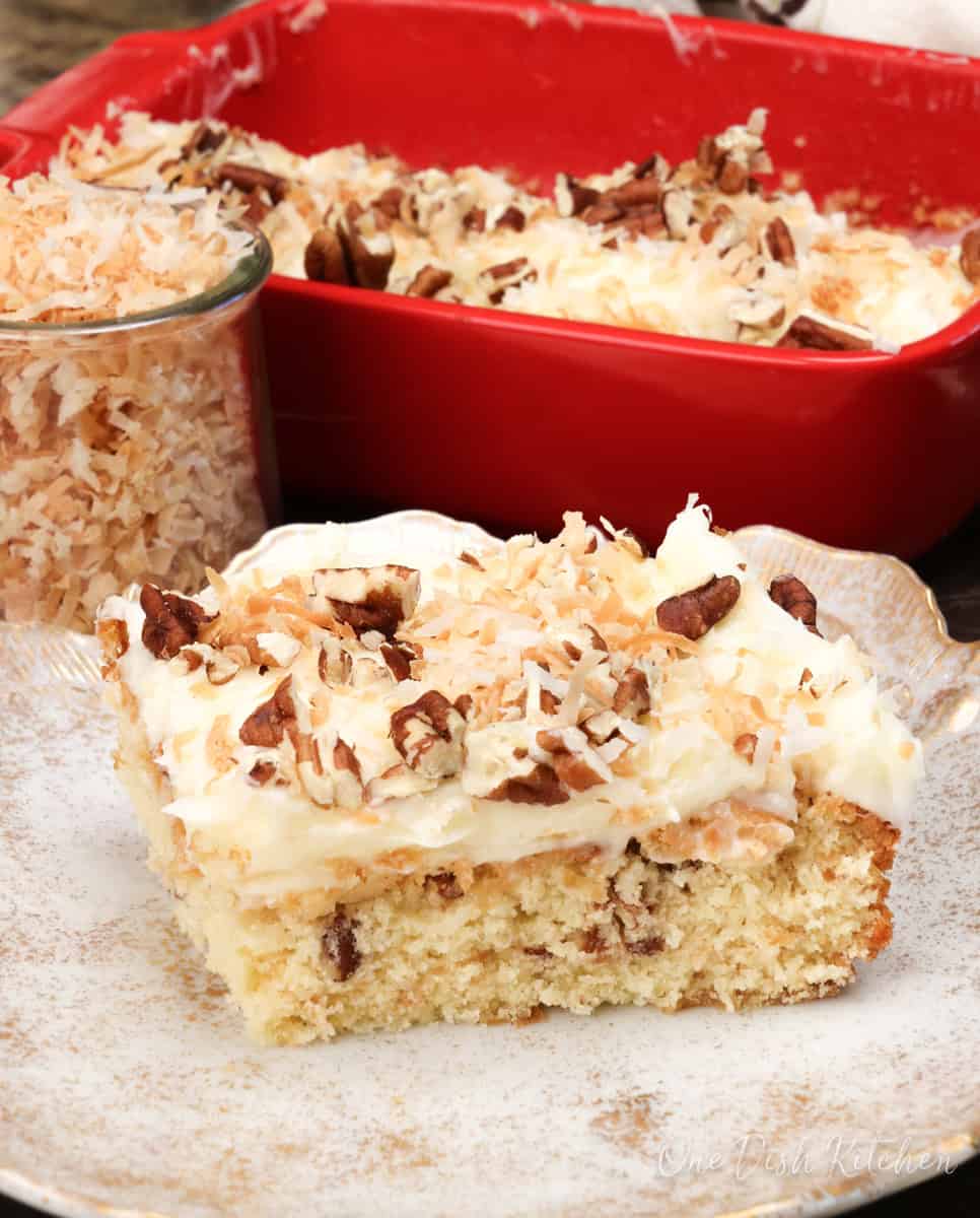 a slice of italian cream cake on a white plate next to a jar of toasted coconut.