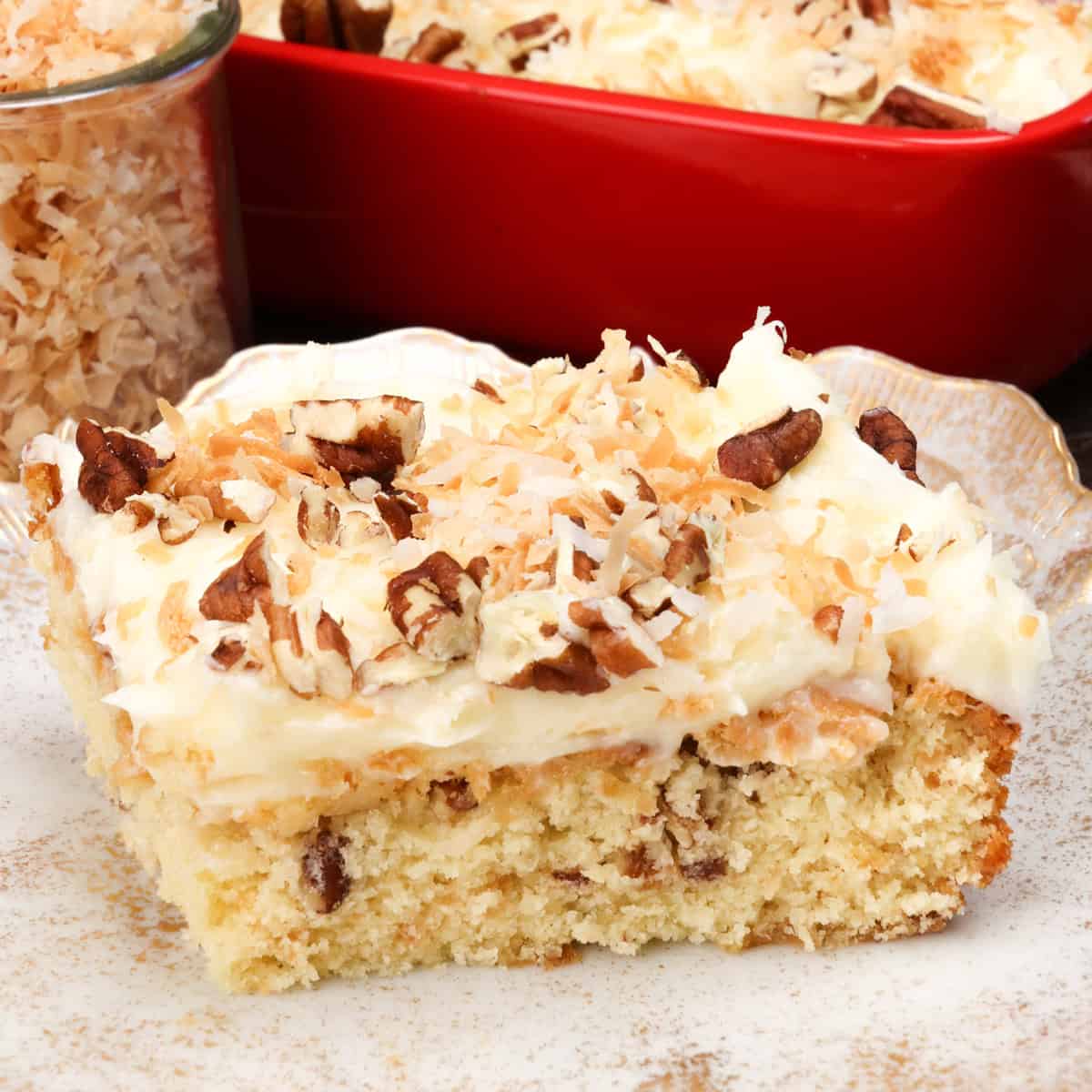 a slice of italian cream cake on a white plate next to a jar of toasted coconut.