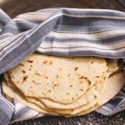 A stack of homemade flour tortillas wrapped in a blue and white striped towel on a metal tray.