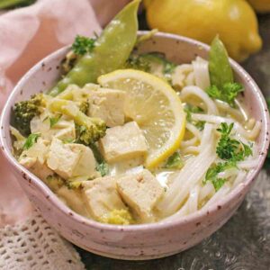 a pink bowl filled with green curry with chunks of chicken and a slice of lemon on top.