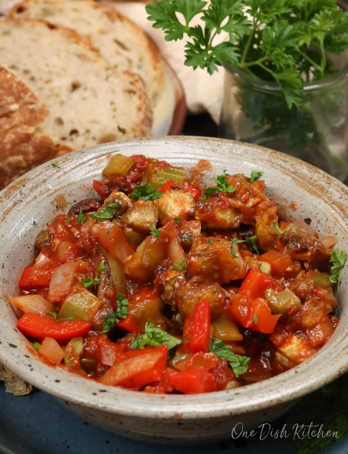 a bowl of caponata next to fresh parsley and slices of bread.