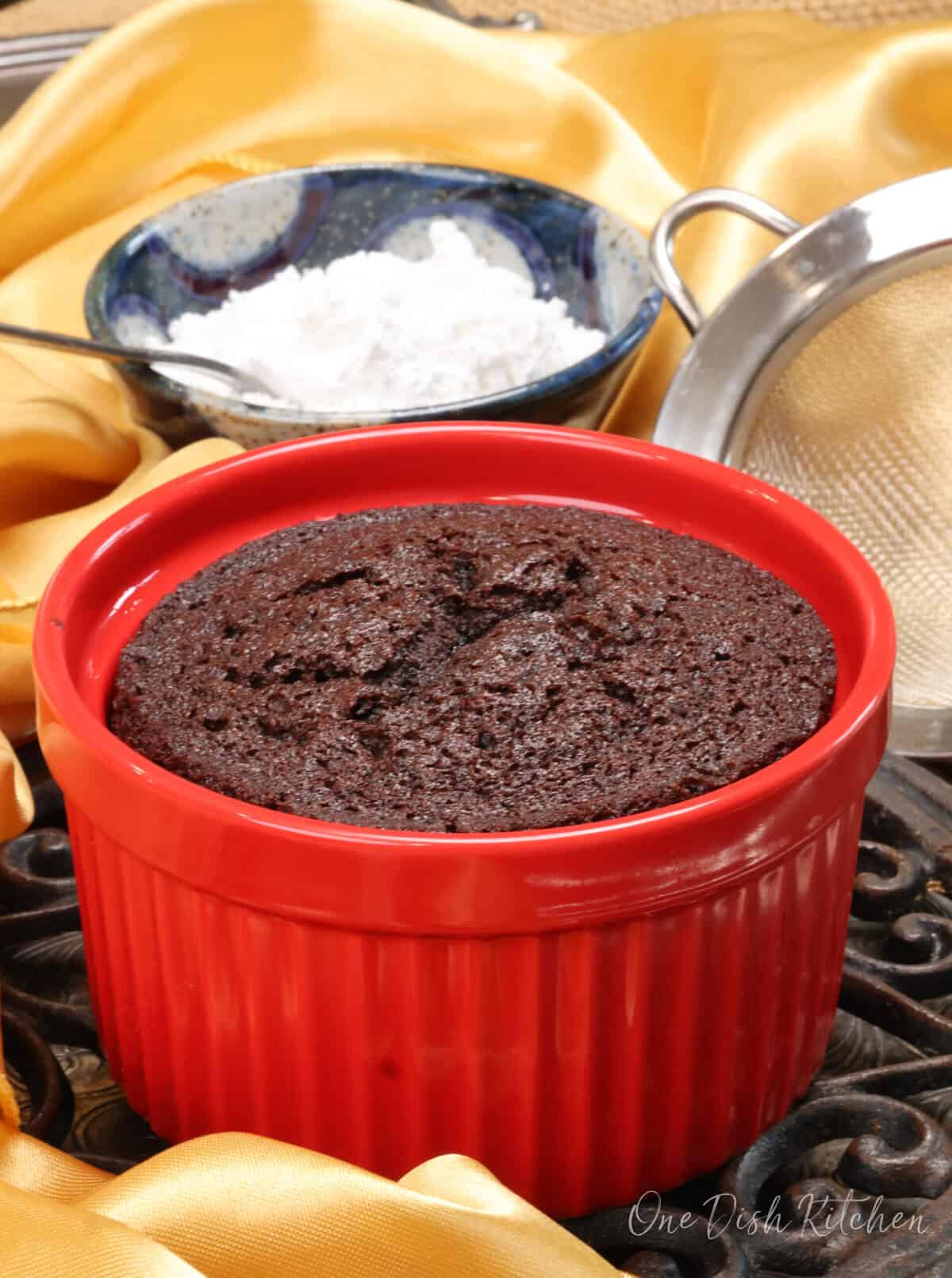 a small chocolate cake cooling on a rack next to a bowl of powdered sugar.