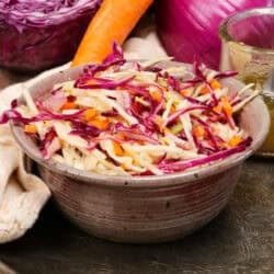 a bowl filled with coleslaw next to a brown napkin, cabbage, onions, and carrots.