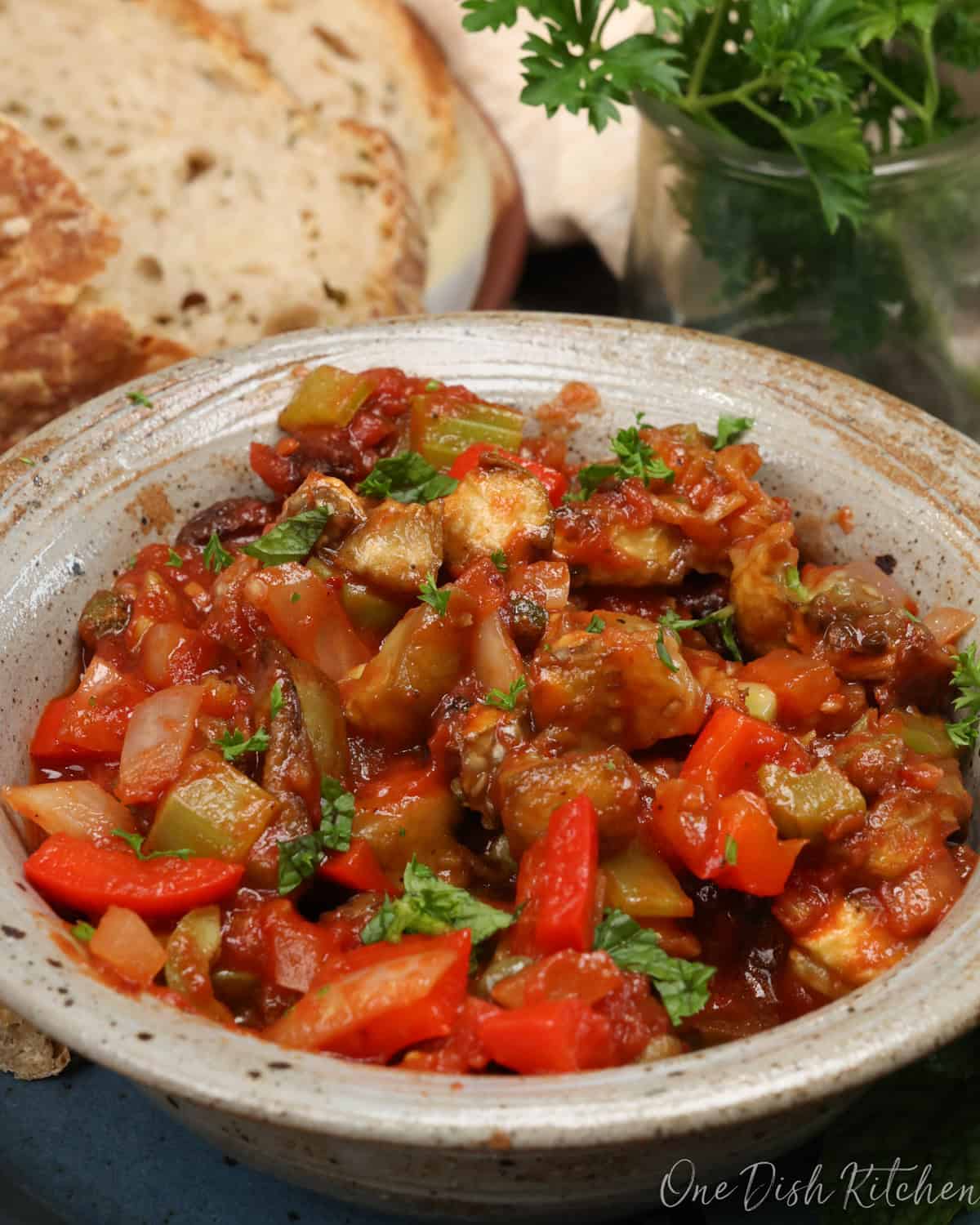 a bowl of caponata next to fresh parsley and slices of bread.