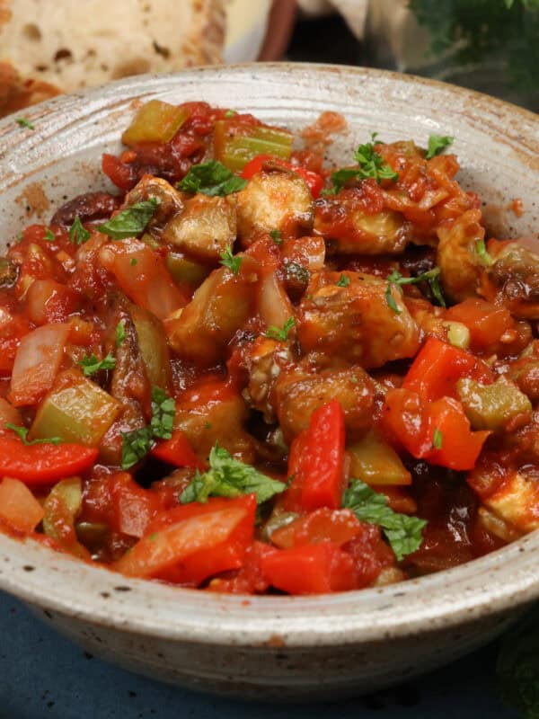 a bowl of caponata next to fresh parsley and slices of bread.